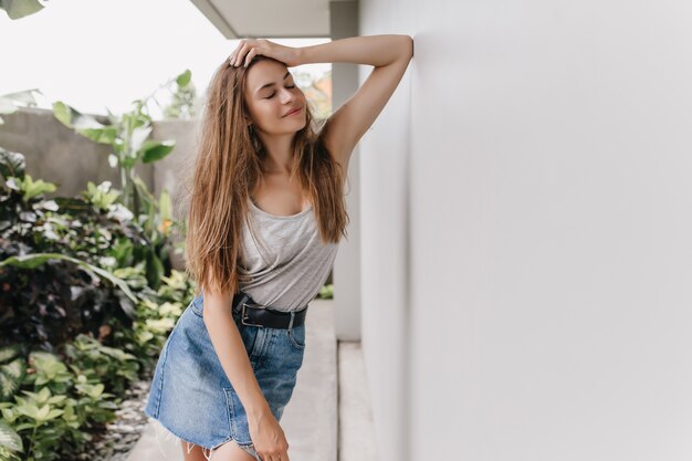 Magnífica mulher de cabelos compridos posando com os olhos fechados perto do arbusto verde. Foto ao ar livre de uma garota relaxada de cabelos castanhos usando saia jeans