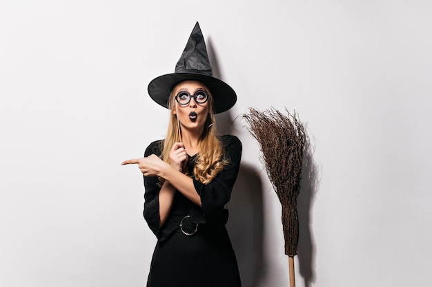 Foto grátis mago espantado de óculos em pé na parede branca. bruxa engraçada emocional posando com chapéu e vassoura.