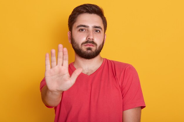 Malm barbudo jovem vestindo camiseta casual vermelha em pé com parada aviso gesto mão e olhando para a câmera com cara séria