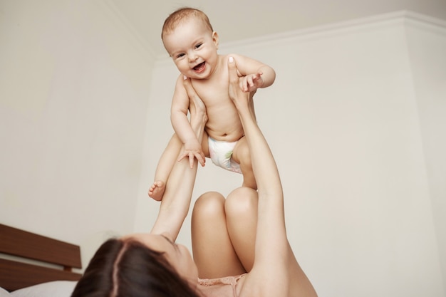 Mamãe linda alegre e seu bebê brincando sorrindo alegremente deitado na cama em casa