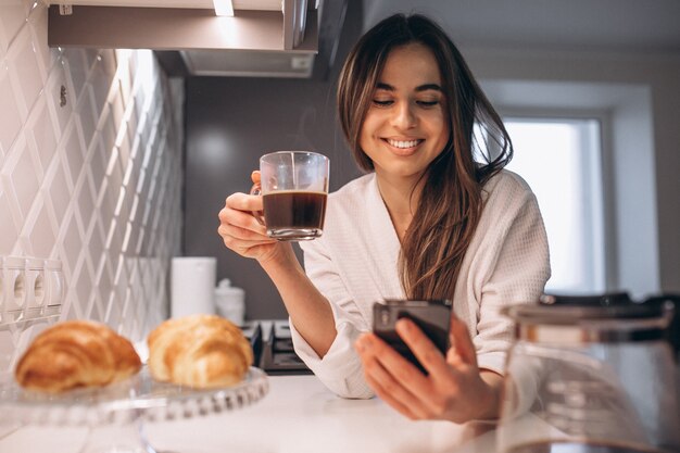 Manhã de mulher com telefone, croissant e café na cozinha