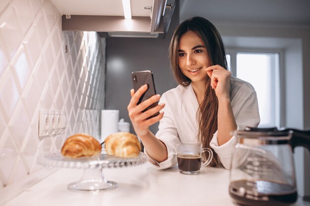 Manhã de mulher com telefone, croissant e café na cozinha