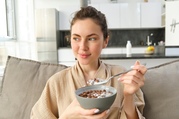 Foto grátis manhã relaxante em casa, uma jovem linda mulher de roupão comendo cereais no sofá com ela