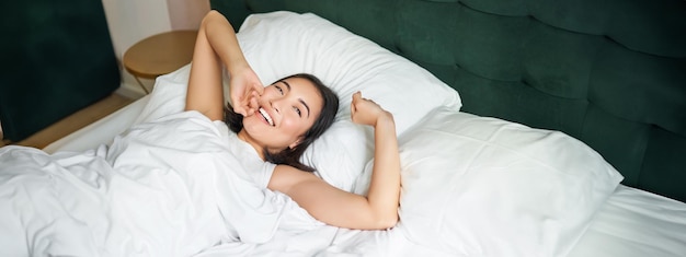 Foto grátis manhãs felizes, linda garota asiática acorda em um terno de hotel, deitada na cama branca, esticando os braços