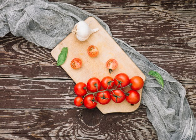 Manjericão; tomate cereja e dentes de alho na tábua com pano cinzento sobre o pano de fundo texturizado de madeira