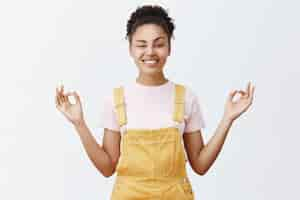 Foto grátis mantenha a calma e livre de estresse com a meditação. mulher afro-americana moderna, alegre e divertida, de macacão amarelo, olhando com um dos olhos e sorrindo abertamente, de mãos dadas em pose zen