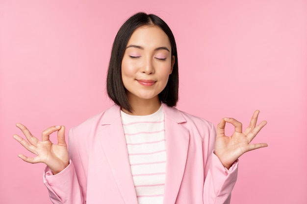 Mantenha a calma Sorridente jovem asiática meditando praticar yoga mindfulness e relaxamento na respiração de trabalho inalar ar com cara satisfeita em pé sobre fundo rosa