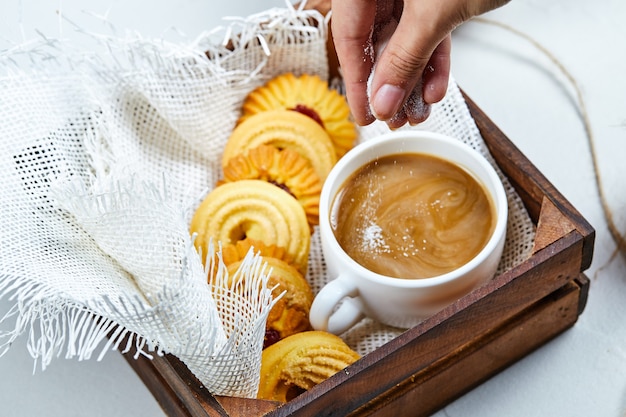 Mão adiciona pó ao café e um prato de biscoitos.