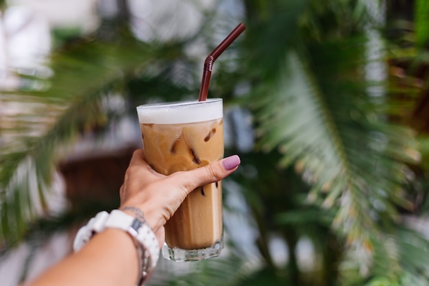 Mão da mulher segurando café com leite gelado sobre arbustos tropicais verdes