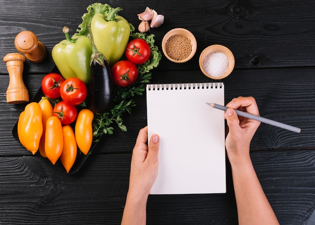 Mão da pessoa, escrevendo no bloco de notas em espiral perto de legumes e especiarias na superfície de madeira preta