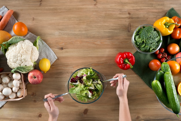 Mão da pessoa preparando fresca salada saudável perto de variedade de legumes e frutas no balcão da cozinha de madeira