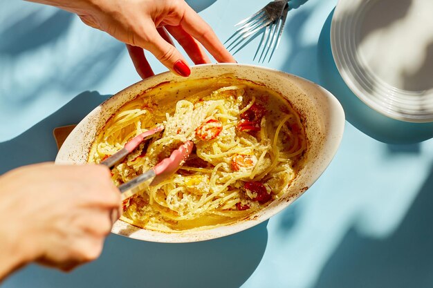 Mão de mulher cozinhando tendências de tomate assado viral e queijo feta com macarrão, fetapasta em uma luz solar de fundo azul, hardlight. prato de jantar novo e popular, vista superior, espaço de cópia.
