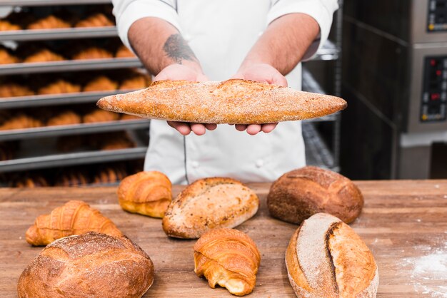 Mão de um padeiro masculino segurando pão baguete por cima da mesa na cozinha comercial