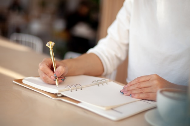 Mão de uma mulher bem preparada segurando uma caneta de ouro e escrevendo notas com uma caneta de ouro no caderno ao lado da janela