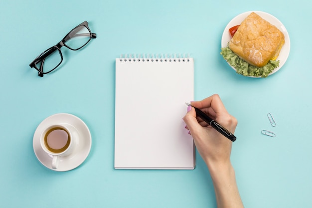 Mão de uma mulher de negócios, escrevendo no bloco de notas em espiral com a xícara de café e sanduíche na mesa do escritório