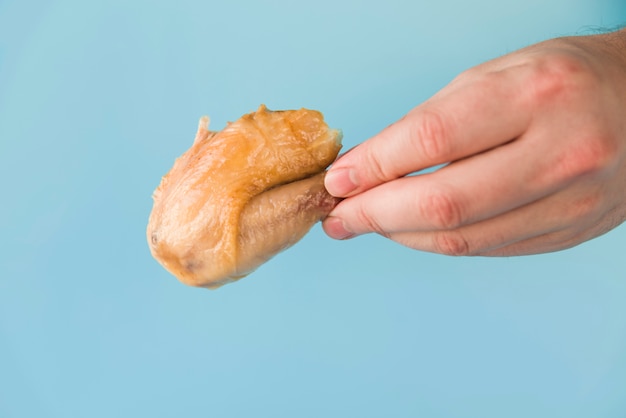 Mão de uma pessoa segurando a perna de frango assada na frente de fundo azul