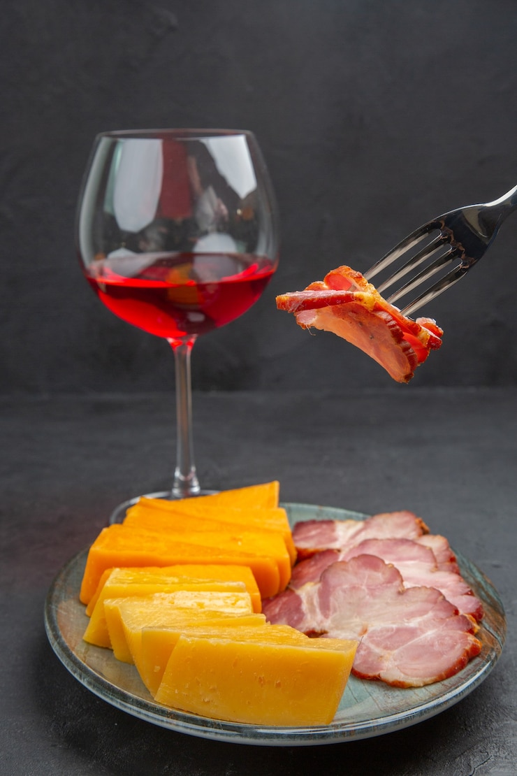Foto grátis mão de visão vertical pegando uma comida com o garfo de um prato azul com deliciosos petiscos e vinho tinto em uma taça de vidro em um fundo preto