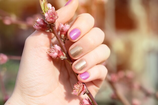 Mão feminina com design de unha ouro e roxo, segurando o ramo de flor.