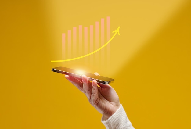 Mão feminina segura um smartphone com gráficos holográficos em um fundo amarelo. crescimento de desempenho, sucesso