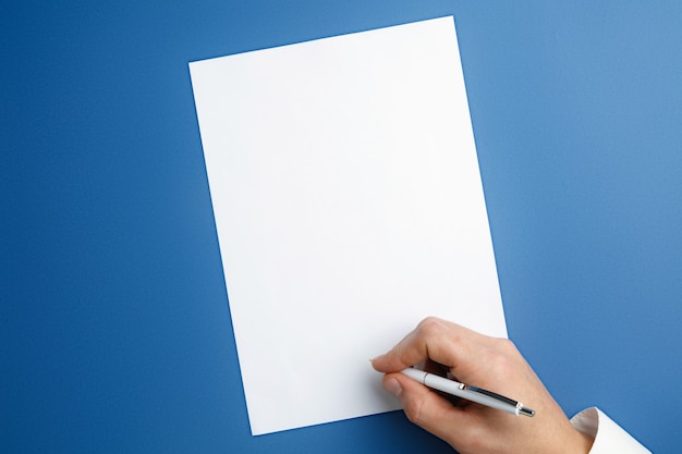 Mão masculina segurando a caneta e escrevendo na folha vazia na parede azul para texto ou desenho. Modelos em branco para contato, publicidade ou uso nos negócios. Finanças, escritório, compras. Copyspace.