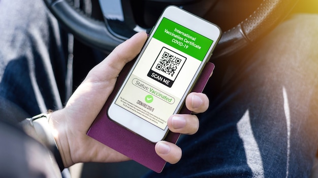 Mão masculina segurando passaporte e smartphone com código QR COVID-19 do Certificado de Vacinação Internacional em um carro