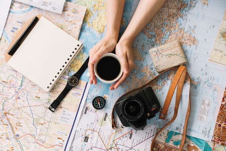 Foto grátis mão segurando a xícara de café no mapa rodeado com o bloco de notas em espiral; bússola; relógio de pulso; câmera e cinta