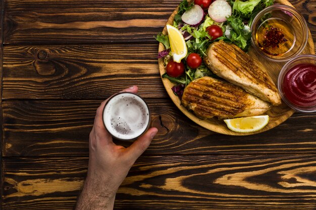 Mão segurando cerveja perto de frango e salada