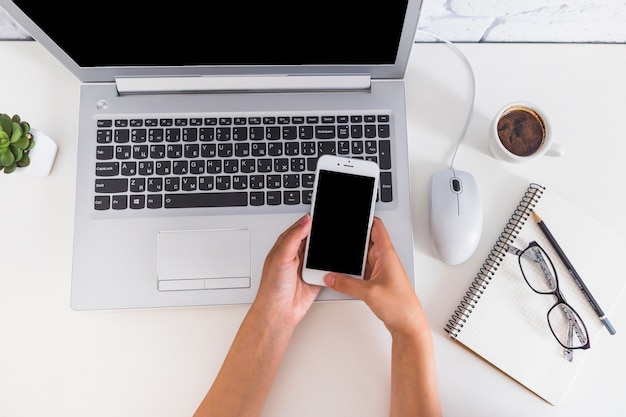 Mão segurando o celular sobre o laptop aberto na mesa