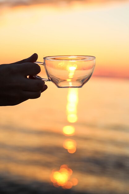 Mão segurando o copo de vidro na frente do pôr do sol