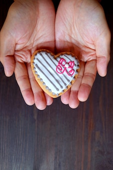 Mão segurando um biscoito com cobertura de chocolate branco em forma de coração em fundo de madeira marrom escuro