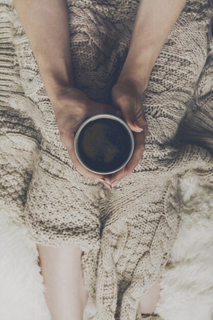 Mãos da mulher segurando café espresso quente saboroso em copo cerâmico sentado na cama com xadrez. Conceito de casa. Vista do topo.