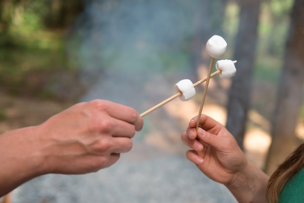 Mãos de casal segurando marshmallows em varas de madeira para churrasco em acampamento na floresta