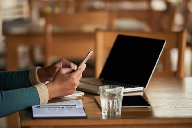 Mãos de homem irreconhecível usando smartphone no café e laptop na mesa