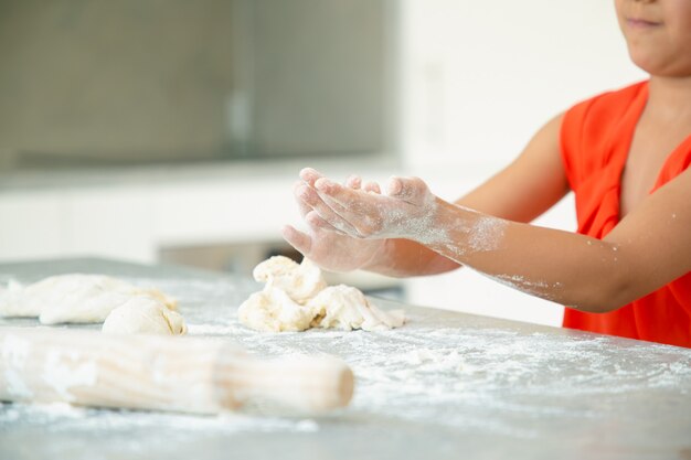 Mãos de menina amassando massa na mesa da cozinha com farinha bagunçada. Criança assando pães ou tortas sozinha. Tiro médio. Conceito de cozinha familiar