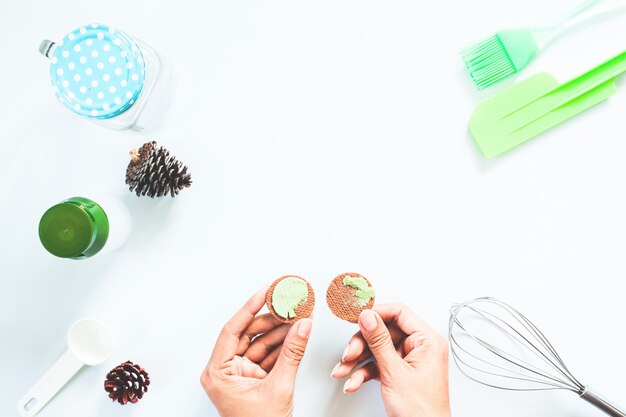 Mãos de mulher com biscoito de creme de chá verde macha e ferramentas de padaria na mesa branca, Flat Lay