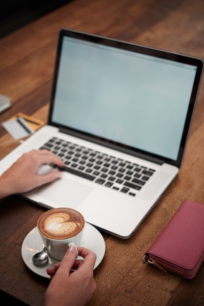 Mãos de mulher irreconhecível com cappuccino e laptop sentado à mesa de madeira