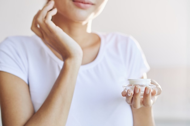 Mãos de mulher irreconhecível, segurando o pote e espalhando o creme para o rosto na pele