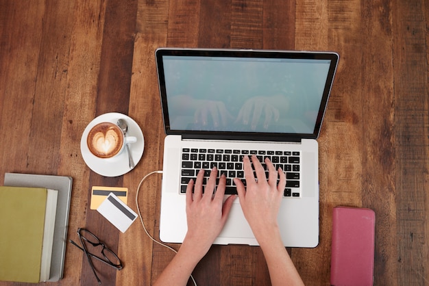 Mãos de mulher irreconhecível, trabalhando no laptop, com cappuccino e cartões de crédito na mesa