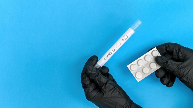 Mãos em luvas médicas pretas segurando o termômetro e a embalagem de comprimidos. Fundo azul. Vista do topo