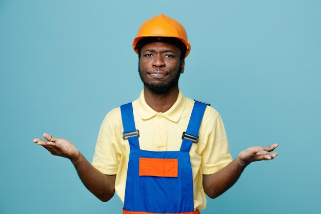 Mãos espalhadas confusas, jovem construtor americano africano de uniforme isolado em fundo azul