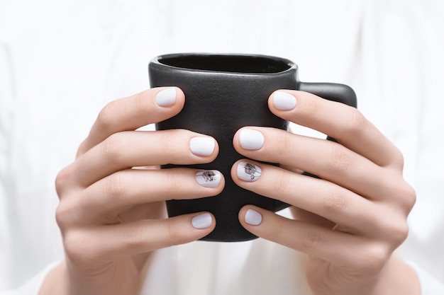 Mãos femininas com design de unhas brancas, segurando o copo preto.