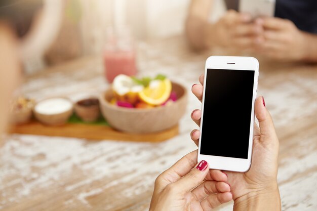 Mãos femininas elegantes com unhas vermelhas limpas, usando telefone celular branco, visualizando feed de notícias através da sua conta nas redes sociais