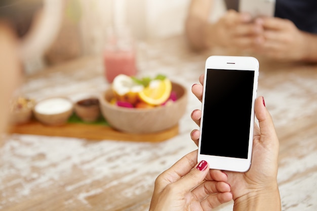 Mãos femininas elegantes com unhas vermelhas limpas, usando telefone celular branco, visualizando feed de notícias através da sua conta nas redes sociais