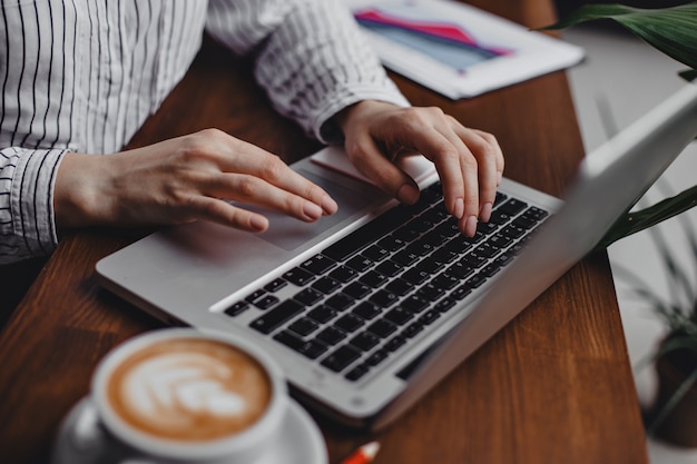 Mãos femininas pressionam as teclas do laptop enquanto está sentado à mesa de madeira com uma xícara de cappuccino branca.