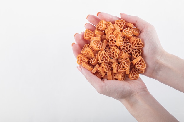 Mãos femininas segurando macarrão em forma de coração cru em branco.