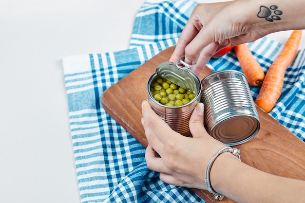 Mãos segurando uma lata de ervilhas cozidas em uma mesa branca com legumes e toalha de mesa.