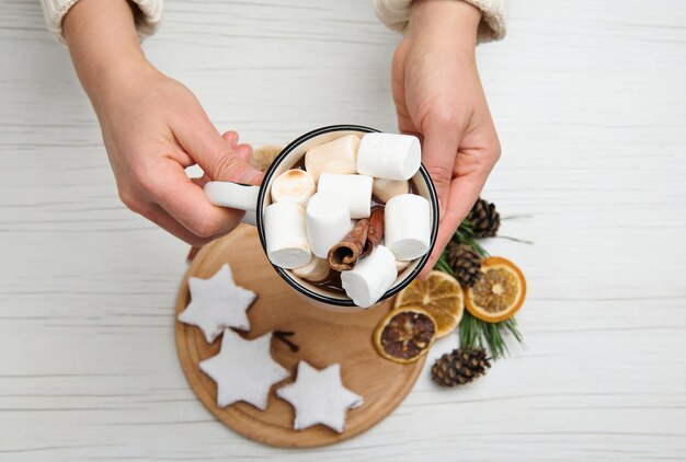 Mãos segurando uma xícara de bebida de chocolate quente com marshmallows sobre uma placa de madeira com biscoitos de gengibre em forma de estrela polvilhados com açúcar de confeiteiro e rodelas de laranja secas ao lado de pinhas e galhos