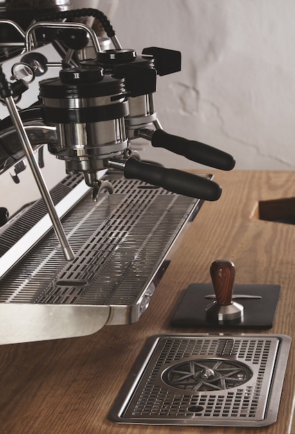 Máquina de café profissional cromada de vista lateral com duas cabeças e portafiltros carregados em cafeteria em mesa grossa de madeira e tamper em couro padespresso, cappuccino, máquina de café com leite.