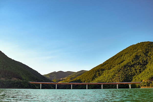 Mar com uma ponte de concreto cercada por colinas cobertas de florestas sob a luz do sol