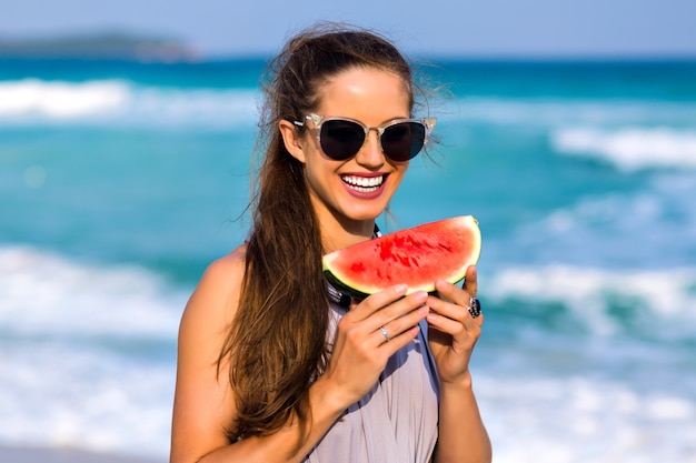Maravilhosa garota de cabelos escuros em óculos de sol, posando no resort do mar nas férias de verão. Retrato ao ar livre do modelo feminino moreno, segurando a melancia e olhando para o fundo do oceano.